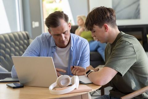 Father tutoring teenage son in living room using laptop and notebook for home learning