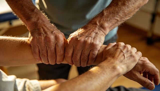 Therapist hands guiding elderly forearm during rehabilitation, caring grip and warm light