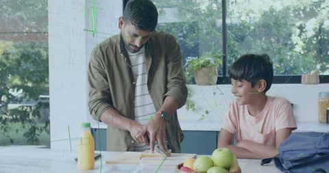 Father slicing bread on sunlit kitchen island while son watching and smiling at breakfast