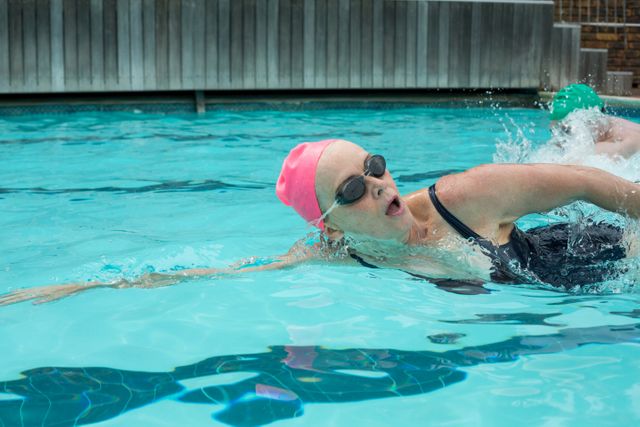 Mature woman swimming in an outdoor pool, wearing a pink swim cap and goggles. Ideal for use in content related to fitness, active lifestyles, healthy aging, sports, and swimming techniques. Perfect for promoting health and wellness programs, senior fitness classes, and swimming lessons.
