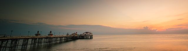 Panoramic view of a wooden pier extending into calm ocean waters at sunset. The sky displays vibrant colors as the sun sets over the horizon. Ideal for travel websites, nature blogs, meditation content, and desktop wallpapers highlighting scenic beauty and tranquility.