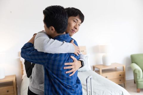 Young asian men embracing in bright bedroom during move with suitcase and backpack