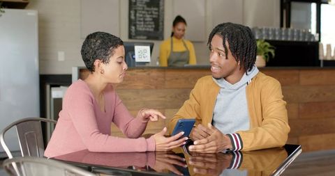 Young friends sharing smartphone at cafe table, woman pointing while man listens