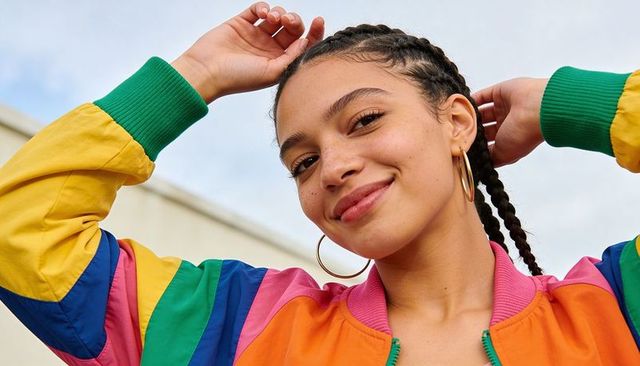 Smiling woman adjusting cornrows wearing colorful bomber jacket and gold hoop earrings