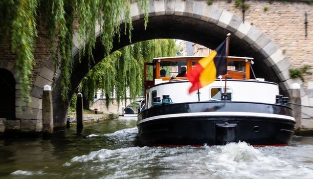 Tour boat passing under stone arch bridge on belgian canal, willow-lined historic quay
