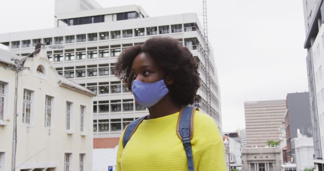 Young African American woman wearing a face mask walking in an urban environment, highlighting contemporary urban life during a pandemic. Useful for content related to city living, health and safety, social narratives, and modern lifestyle.