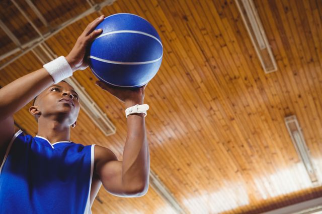 Teenage boy in blue jersey preparing to shoot basketball in an indoor court with wooden ceiling. Ideal for use in sports-related content, youth athletic programs, basketball training materials, and motivational posters.