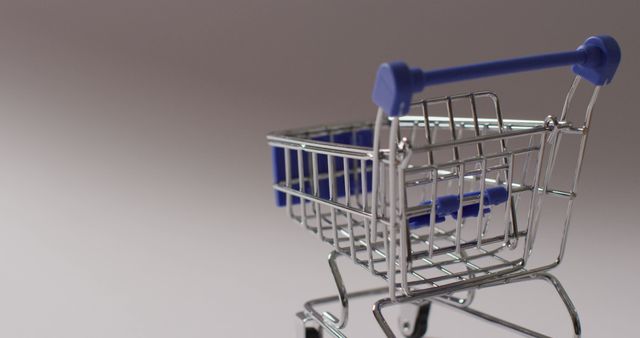 Miniature shopping cart with blue handle seen in close-up against neutral background. Perfect for illustrating concepts related to retail, online shopping, e-commerce, and consumerism. Suitable for use in advertisements, promotional materials, or as a visual aid in presentations about shopping and economic trends.