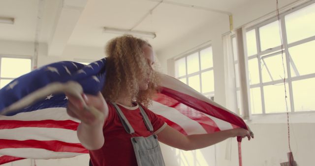This image shows a young woman with curly hair joyfully waving an American flag indoors. She is wearing casual clothes and appears to be celebrating. Perfect for themes related to patriotism, national pride, Independence Day, or American culture.