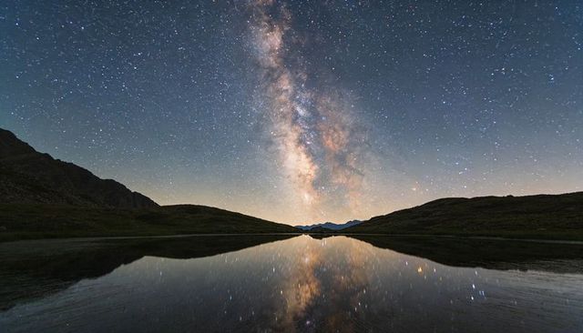 Milky way reflecting in calm alpine lake with starry sky and mountain ridge