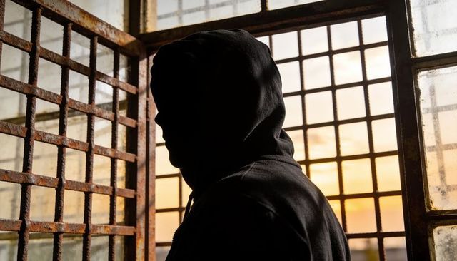 Silhouetted hooded figure standing by rusted barred window at sunset