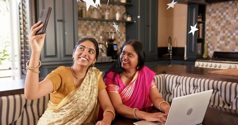 Indian mother and daughter sharing joyful tech moment in bright kitchen wearing sarees