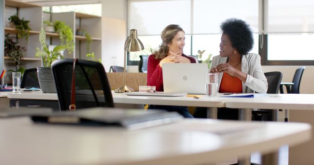 Two businesswomen having a discussion at an office desk, highlighting collaboration and teamwork. Ideal for depicting corporate environments, partnership, and project planning.