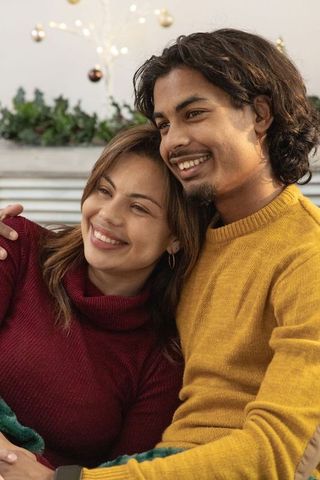Smiling couple cuddling on couch wearing cozy sweaters, warm holiday living room