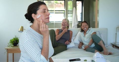 Mature women enjoying self-care at home using jade roller and face masks on sunlit bed