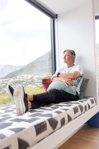 Mature man relaxing on window bench hugging cushion with scenic mountain valley view