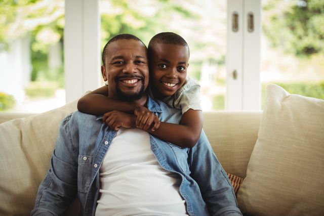 This image captures a joyful moment between a father and his son, with the son embracing his father while they sit on a couch at home. Perfect for use in family-oriented advertisements, parenting blogs, lifestyle articles, and promotional materials that emphasize family bonds and happiness.