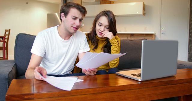 A young couple sits on a sofa in home, actively reviewing financial documents together. They appear focused and thoughtful, concentrating on the papers spread out before them, laptop seen next to them on a wooden table, suggesting modern lifestyle and digital integration. This can be used in articles or ads related to financial planning, adult education, home budgeting, mortgage planning, or domestic life.