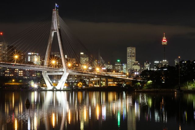 This image showcases Sydney's Anzac Bridge and city skyline illuminated at night. The bridge and buildings reflect beautifully on the water, creating a vibrant urban night scene. Ideal for travel blogs, architectural designs, wallpapers, and articles highlighting Sydney’s landmarks.