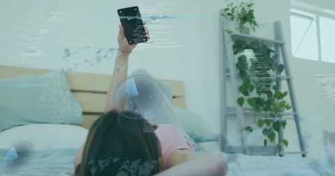 Korean woman lying on bed raising smartphone with futuristic hud in pastel bedroom