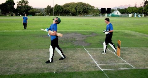Batsmen walking between wickets on cricket pitch wearing blue shirts and protective gear