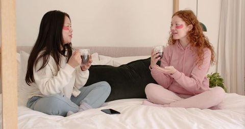 Two friends wearing under-eye patches sipping warm drinks on bed in cozy loungewear, chatting