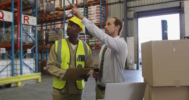 Front view of a middle aged Caucasian male warehouse manager holding a barcode scanner and a young African American male warehouse worker holding a clipboard standing by a stack of boxes in a warehouse loading bay talking, looking at a laptop computer, pointing and shaking hands. They are working in a freight transportation and distribution warehouse. Industrial and industrial workers concept 4k