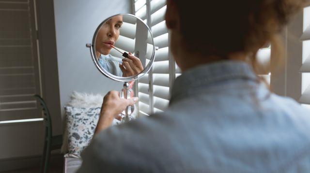 Woman sitting by window applying lipstick using handheld mirror. Ideal for beauty, self-care, and lifestyle content. Highlights natural light and cozy home environment, perfect for promoting makeup products or home decor.