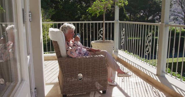 Elderly woman enjoying morning coffee while sitting on wicker chair on balcony. Sunlight casting warm shadows creates a peaceful, relaxing environment. Ideal for use in articles on retirement, healthy living, leisure activities for seniors, and advertisements promoting senior living communities.