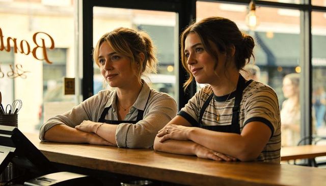 Leaning and chatting baristas at sunlit cafe counter with pos, aprons, cozy urban vibe