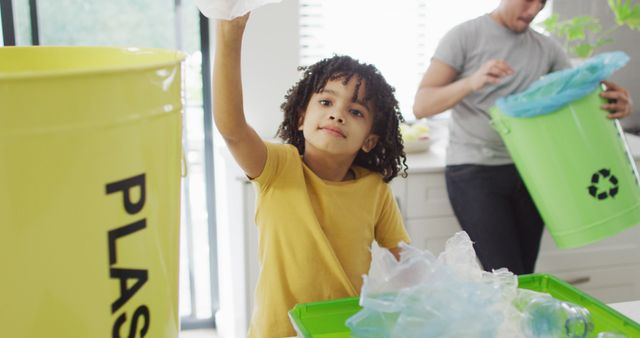 Child placing plastic waste into appropriate recycling bin in modern kitchen while parent assists with sorting. This can be used for environmental awareness campaigns, educational materials on recycling, family activities promoting green living, and sustainability initiatives.