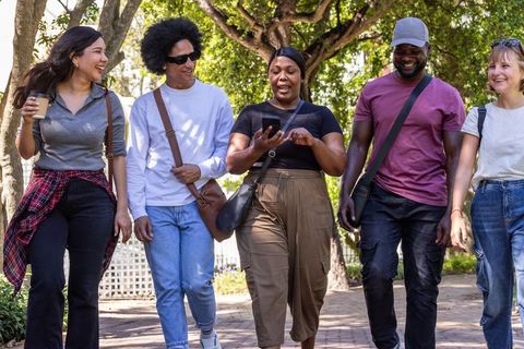 Multicultural friends walking in leafy park laughing, chatting and sharing smartphone