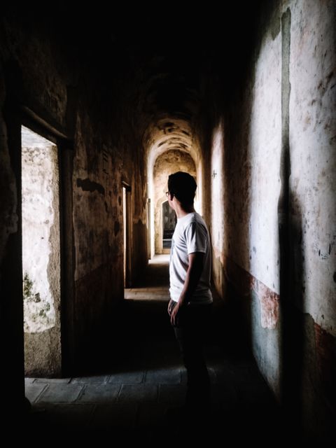 This image depicts a man standing in an abandoned corridor with sunlight streaming through a door, creating a moody and mysterious scene. Perfect for themes related to exploration, urban decay, solitude, adventure, and architectural photography. It can be used in articles, blogs, and promotional materials focused on adventure, mystery, or exploring forgotten places.