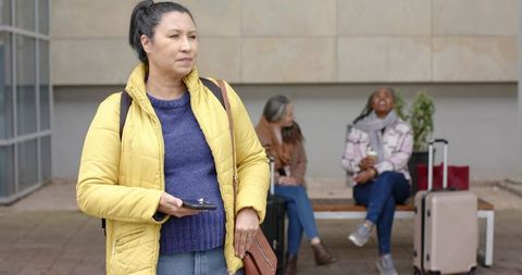 Mature traveler in yellow puffer coat standing with smartphone and suitcase at station bench