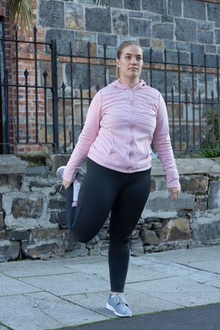 Young woman stretching quads on urban sidewalk wearing pink jacket and athletic leggings