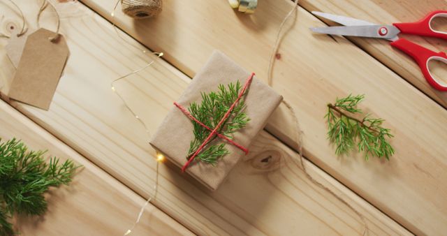 Gift wrapped in brown paper and red string, adorned with pine branches, on light wooden table. Small scissors, jute twine, and blank gift tag nearby. Ideal for holiday marketing, DIY project tutorials, Christmas crafts, festive articles, or ads focused on gifting and wrapping ideas.