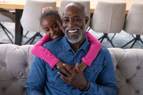 Grandfather and granddaughter sharing joyful hug on sofa, multigenerational family bonding