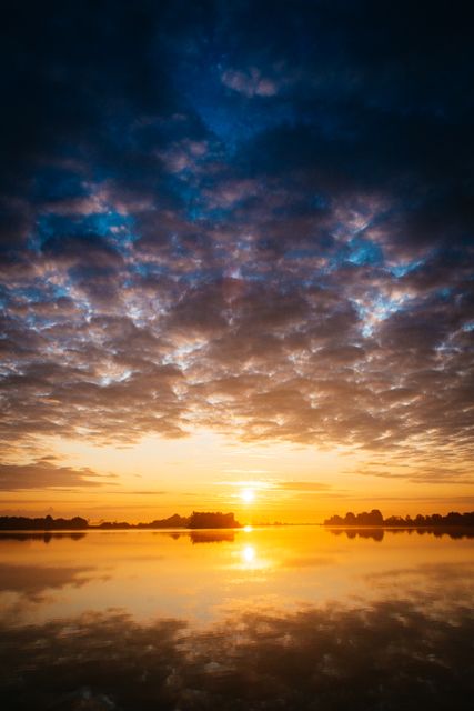 Capturing a stunning sunrise over a tranquil lake with perfect reflections on the water. Vibrant colors and dramatic clouds add to the scenic beauty. Ideal for use in travel magazines, nature calendars, inspirational posters, or desktop wallpapers, promoting peace and natural beauty.