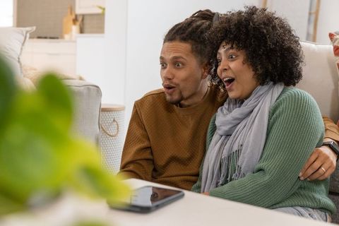 African american couple reacting while watching tablet, cuddling on cozy modern living room couch
