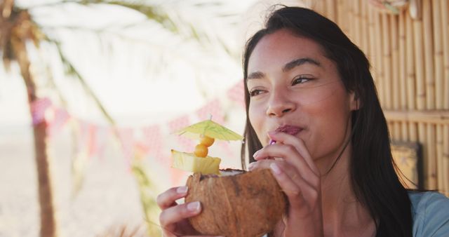 Smiling biracial woman drinking cocktail from coconut shell at beach bar. Summer, free time, relaxation and vacations.