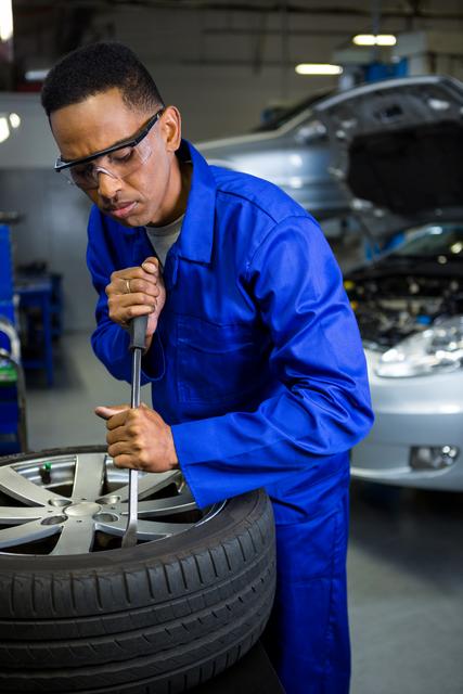 Mechanic in blue uniform and safety glasses repairing a car tire in a garage. Ideal for use in automotive repair advertisements, maintenance service promotions, and educational materials on vehicle care.