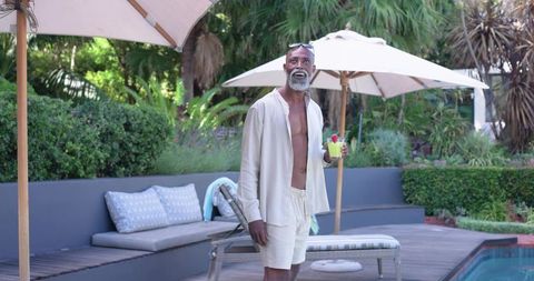 Mature african man standing by pool holding tropical cocktail enjoying sunny patio