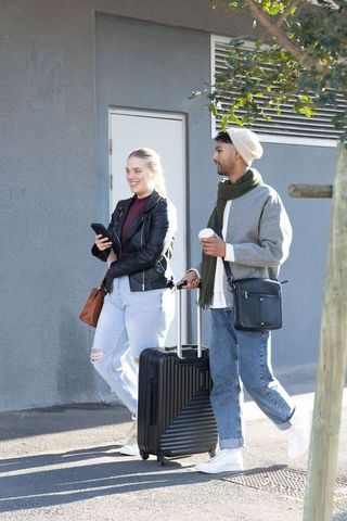 Young couple walking with suitcase and coffee on sunny urban sidewalk, travel lifestyle