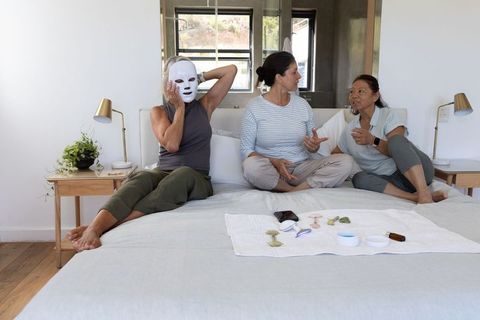 Diverse women sitting on bed wearing led facial mask and sharing home skincare ritual