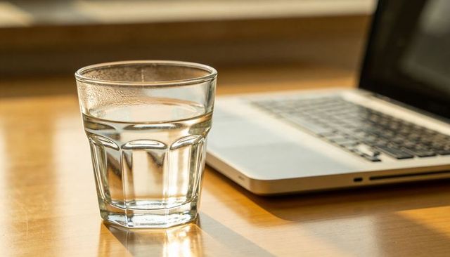 Faceted glass of water casting warm reflection on wooden desk beside laptop in morning sunlight