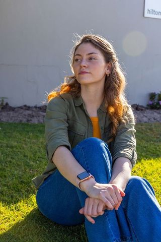 Contemplative young woman sitting on grass backlit by sun wearing smartwatch and denim olive overshi
