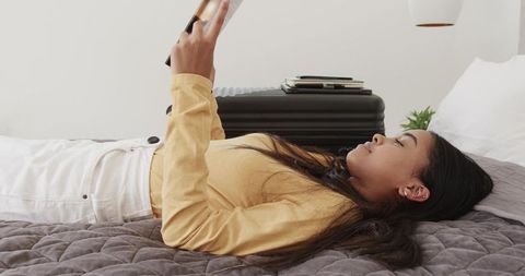 African american woman lying on bed reading paperback near packed suitcase