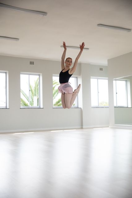 Caucasian female ballet dancer practicing in a bright studio, jumping with arms raised. Ideal for use in articles or advertisements related to dance training, ballet classes, fitness, and performing arts. Can also be used for promoting dance schools, ballet workshops, and physical fitness programs.