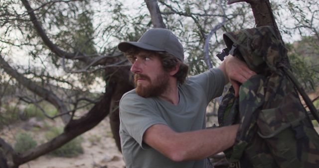 Man with beard wearing hat and t-shirt, holding camouflage jacket in forested area. Suggests themes of adventure, survival, exploration. Ideal for content related to wilderness experiences, camping, outdoor gear promotion, adventure lifestyle.