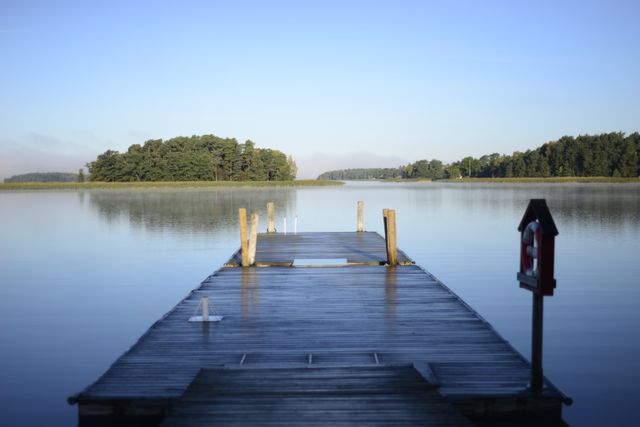 A wooden pier extends into a tranquil lake on a calm morning. The water is still, reflecting the surrounding trees and sky. This scenic outdoor setting is ideal for themes of peace, nature, relaxation, and contemplation. Perfect for use in travel ads, relaxation posters, and nature-themed website backgrounds.